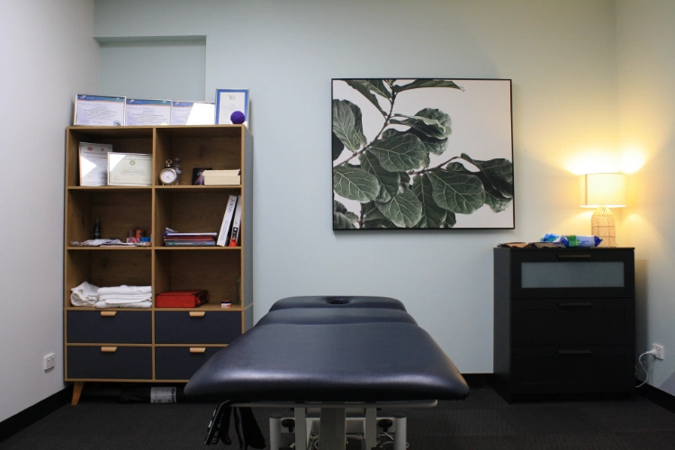 Treatment room at Perth Lymphoedema Clinic with a massage table, shelving unit, and decorative wall art